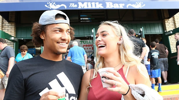 A smiling young couple stands outside "The Milk Bar" building holding small paper cups, laughing together at the Great New York State Fair in Syracuse.