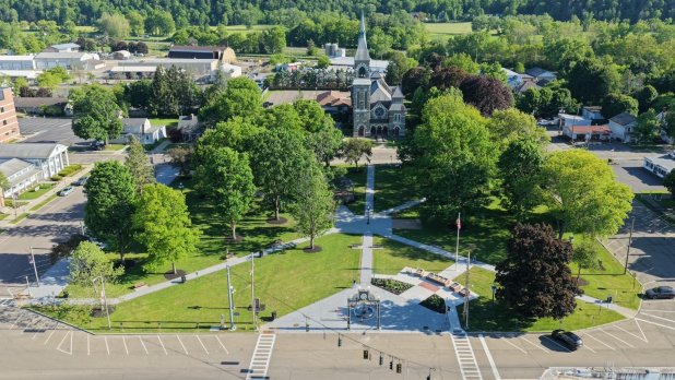 Aerial view of Pulteney Square in Bath, NY, a leafy town park with pathways, flags, and a church centered beyond the green, conveying a peaceful community setting.