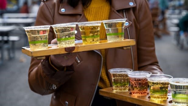 A person carries two cider tasting flights each bearing four plastic cups of various ciders at Bad Seed Cider Company in Highland, New York.