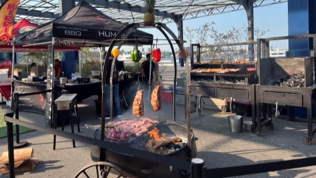 Outdoor barbecue setup with meat and vegetables roasting over an open flame at the Latin Food Fest in New York City. Tents in the background read "Latin American BBQ."