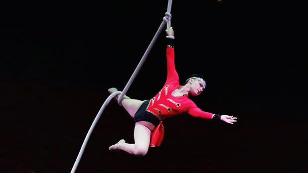 An aerial acrobat performing mid-air while suspended by a thick white rope against a dark background. She wears a long-sleeved red leotard with gold trim, black bottoms, and a sparkling headband, holding the rope with one hand while extending her other arm outward.