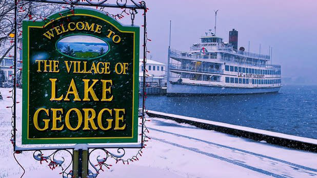 Exterior of a green sign that says "Welcome to the Village of Lake George" with a steamboat in the background covered in snow