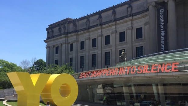 Large yellow letters spelling "YO" in front of the exterior of the Brooklyn Museum with a quote in orange lettering that reads "Do not disappear into silence"