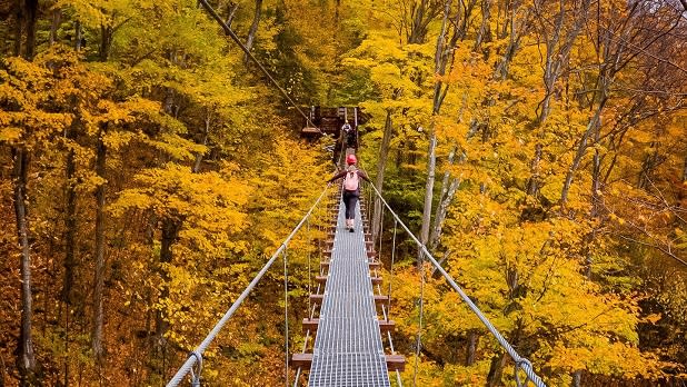 A woman crosses a bridge surrounded by bright yellow foliage at New York Zipline Adventures at Hunter Mountain