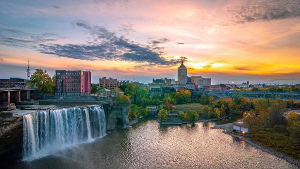 Aerial view of Rochester skyline featuring High Falls, green trees, and tall buildings at sunset