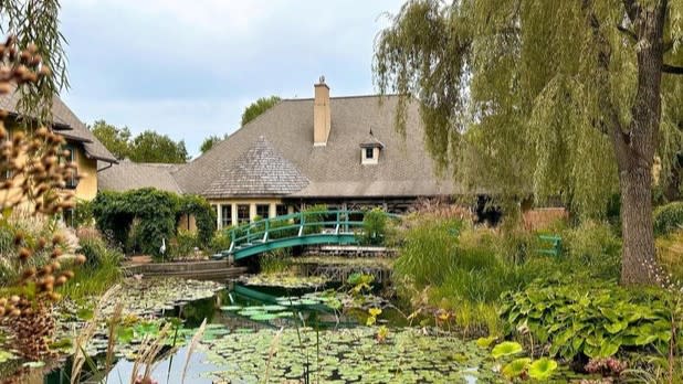 A dark green bridge arches over a pond filled with green lily pads, surrounded by rich green vegetation in front of the exterior of the Mirbeau Inn & Spa