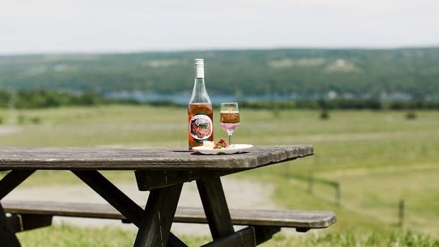 A bottle and glass of rose wine sit on a wooden picnic table in front of a green and blue lakeside view