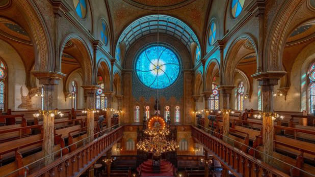 A blue stained glass window illuminating a synagogue with carved walnut balconies, seating, and a chandelier