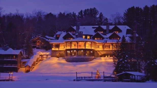 The warmly lit Lake Placid Lodge sits in a snowy landscape at dusk. Soft golden lights create a cozy glow, contrasting with the deep blue sky and snow.