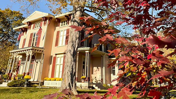 Sunlit historic yellow house with wraparound porch and U.S. flags, framed by vivid red autumn leaves and mature trees; warm, peaceful fall scene.
