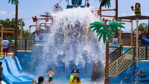 A giant water bucket dumps water on kids playing in the LEGOLAND Water Playground, surrounded by life-sized Lego statues.
