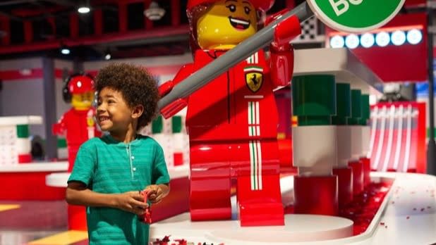a little boy in a turquoise shirt plays with Legos at the LEGO Ferrari Build & Race attraction. Behind him a giant Lego figure dressed in a red Ferrari outfit holds a green sign that says "Go Build"