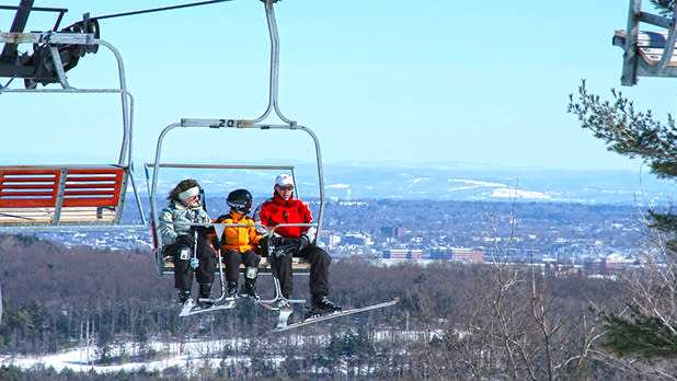 A man, woman and child are sitting on a ski lift traveling along Maple Ridge with a view of the town and snowy hills in the distance