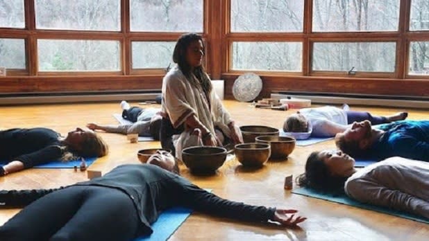Six people lay on their backs with their eyes closed around a women sitting up guiding a meditation