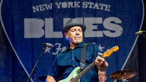 Man playing a light green guitar in a blue shirt and grey hat on stage at the NYS Blues Festival
