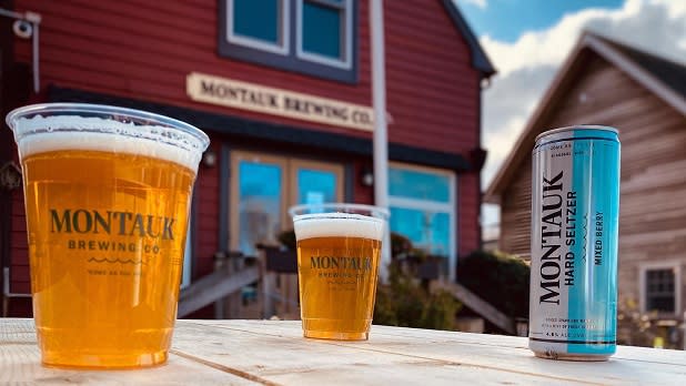 Two cups filled with golden beer and a can of hard seltzer stand on a table outside Montauk Brewing Company