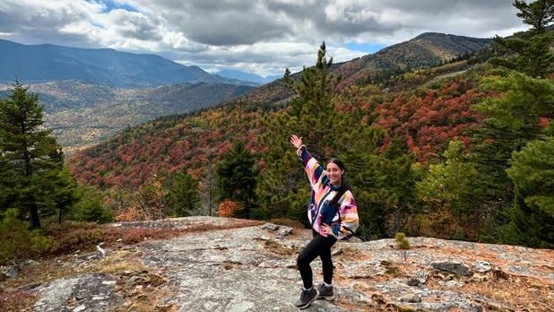 A woman stands a the top of Bear Den Mountain behind a sprawling view of the Adirondack Mountains spotted with orange and red foliage