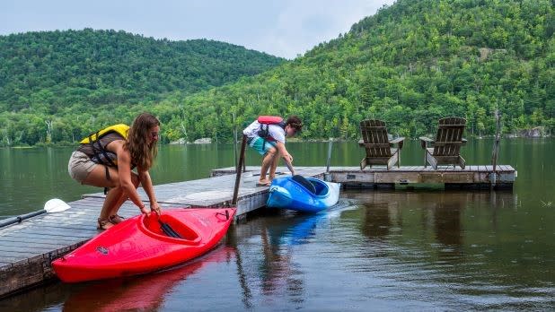 A mother and son placing a red and blue kayak into a lake from a dock surrounded by lush, green mountains