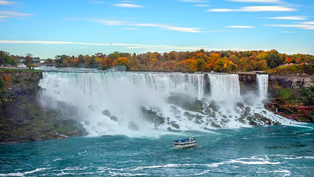 Water flowing down Niagara Falls with the Maid of the Mist boat steering in the direction of the falls below with orange, yellow, and green trees at the top in the distance