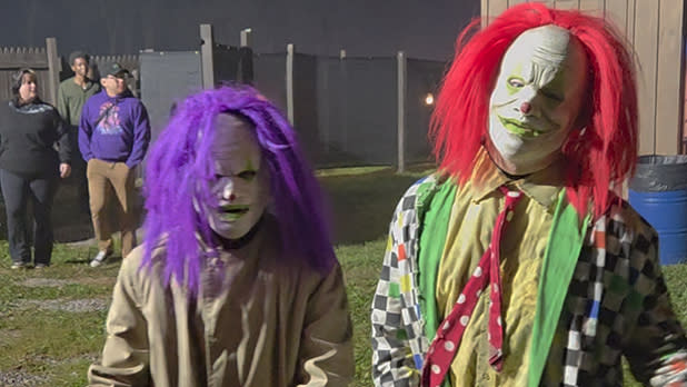 Two costumed clowns pose outdoors at The Haunted Forests in Clarence, NY. One wears a messy purple wig and beige clothes; the other has a bright red wig, white face paint, and a colorful suit.