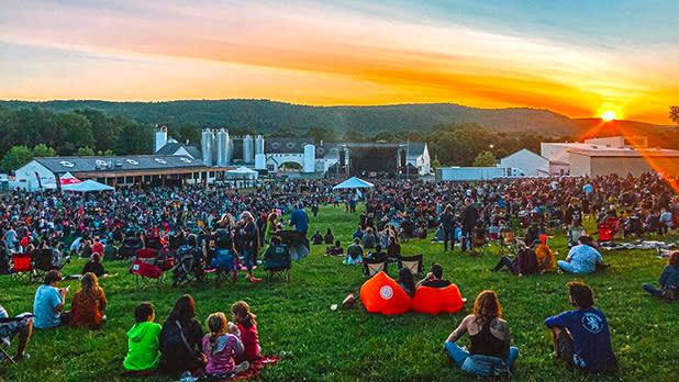 A crowd of people sitting in the field at Brewery Ommegang with the sun setting to the right behind the mountains