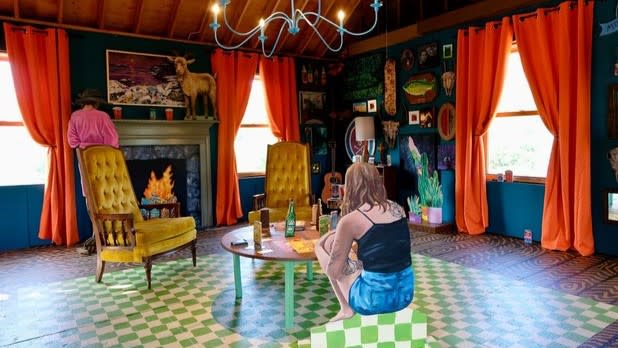 An exhibit at the Wassaic Project of a woman sitting on a green and white checkered floor across the table from yellow chairs.