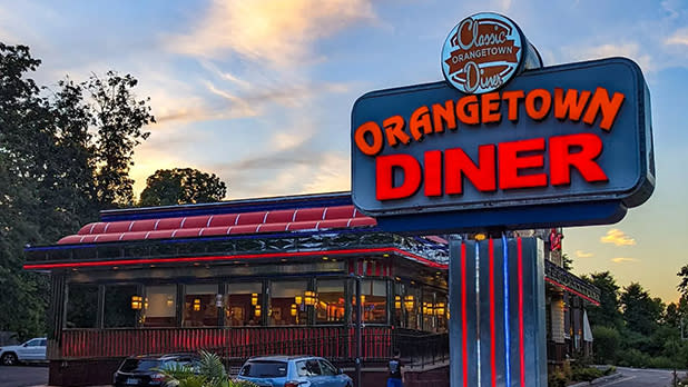 Exterior of Orangetown Diner at dusk with neon sign lit. Trees in the background and a few cars parked in front.