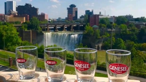 Four tasting glasses that read "Genesee Beer" with a view of High Falls and the Rochester Skyline in the background on a sunny day