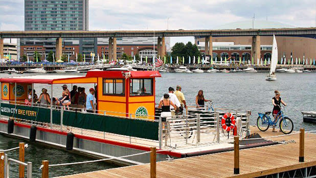 A yellow, green, and red ferry floating at a dock with a person bringing their bike onto the ferry and other people standing on the boat with a big building in the distance