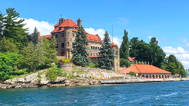 The exterior of Singer Castle on Dark Island from the water surrounded by green trees with a few clouds in the sky