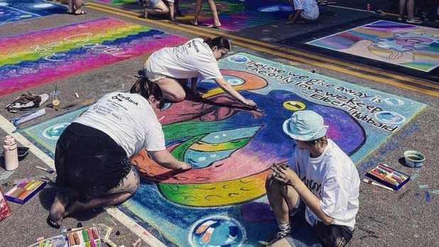 Three people in white t-shirts color a vibrant mural with side walk chalk