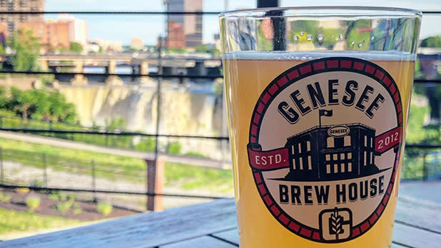 A pint glass labeled with the Genesee Brew House logo on a table with High Falls seen in the background in Rochester. Credit: @roc.beer on Instagram.