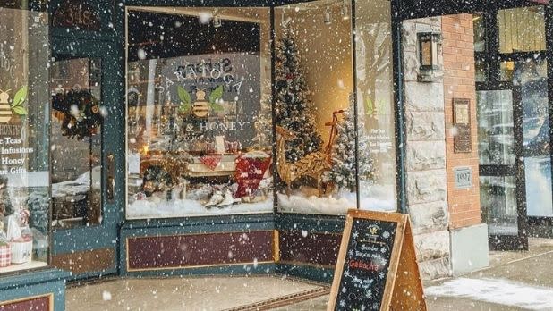 Snow falling in front of the Saratoga Tea & Honey Company store with christmas displays seen through the glass windows