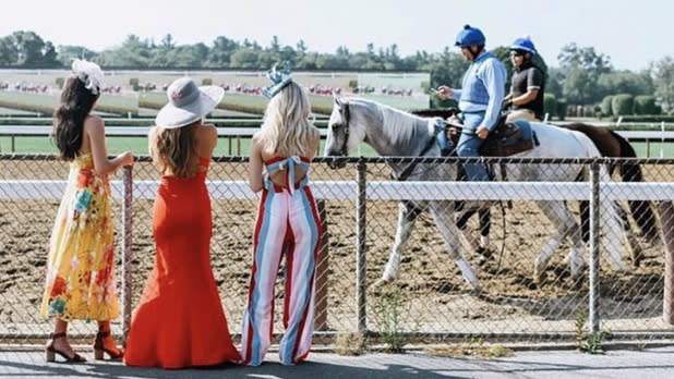 Three women in fancy clothing stand at the fence as two horse and jockeys walk past