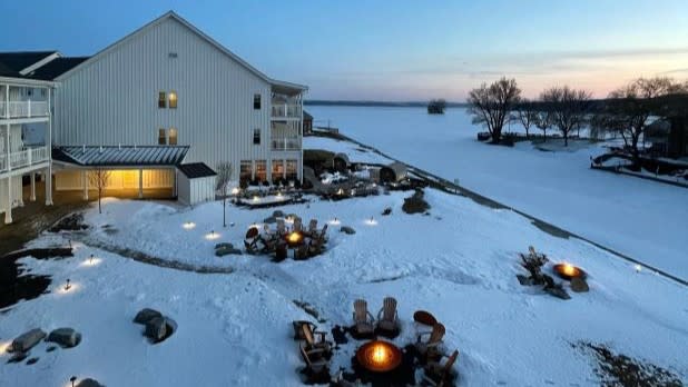 Snow-covered courtyard outside the Lake House on Canandaigua hotel at dusk with lit fire pits surrounded by chairs next to a frozen lake in the background.