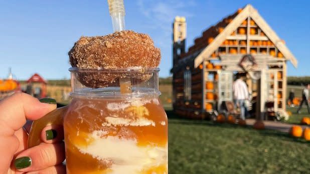 Person holding a "fall shake up" (apple cider drink topped with a cider donut) in front of a house made from pumpkins