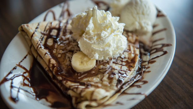 A crepe topped with chocolate drizzle, banana, and whipped cream with vanilla ice cream on the side.