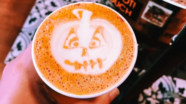 Close-up of a hand holding a foamy latte topped with playful jack-o’-lantern art, evoking a cozy, festive autumn mood at Coffee Labs Roasters in Tarrytown.