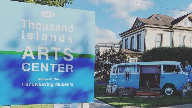 A light and dark blue sign that reads "Thousand Islands Arts Center, home of the Handweaving Museum"
