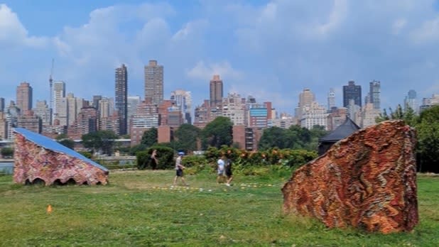 Two orange large half rock sculptures on green grass with a backdrop of the city skyline on a sunny day