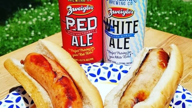 Two Rochester White Hots, hot dogs on a paper plate in front of two cans of Zweigles red and white ale