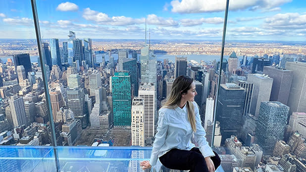 A woman in a white shirt and black pants is sitting on the glass floor by glass walls in the Summit One Vanderbilt building looking out at the city skyline