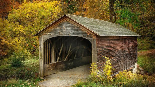 A weathered wooden covered bridge sits among the yellow and orange fall leaves