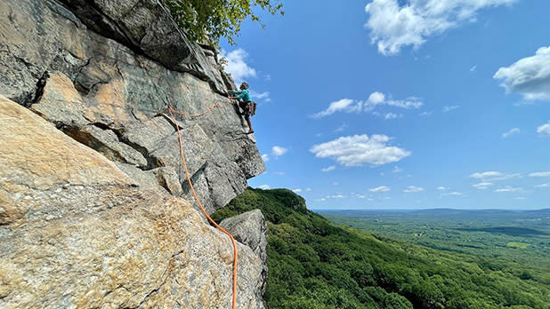 A person in a blue sweater rock climbing a mountain in the 'Gunks overlooking the green forest below