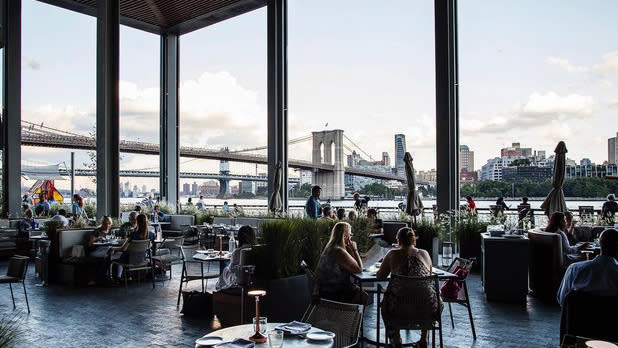 People dine outdoors with view of the East River and the Brooklyn Bridge