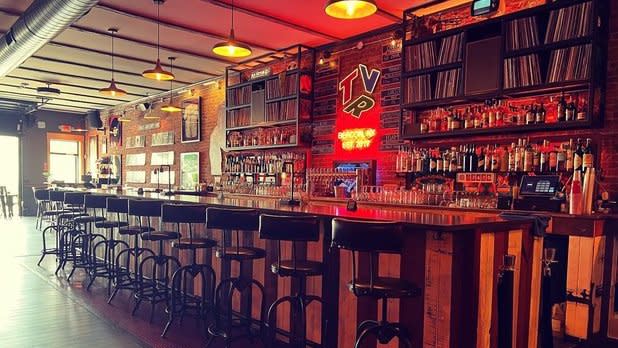 The bar at the Vinyl Room in Beacon, NY lined with bar stools. Shelves of records hang from the back wall that's illuminated by a red neon sign