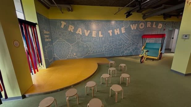 A blue wall with the words "travel the world" written in white, small beige stools sit in front of a small yellow stage next to an exhibit where a bike pulls a carriage.