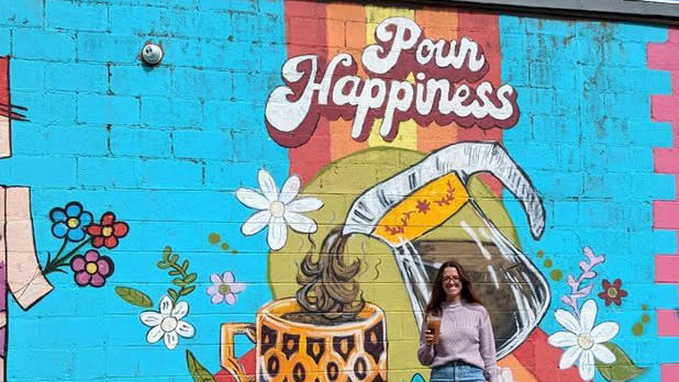 A woman with a beverage stands in front of a bright blue mural that says "pour happiness" above a coffee pot pouring coffee into a yellow and brown mug.