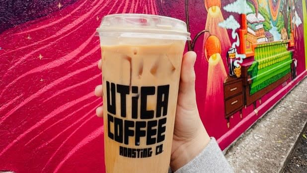 A hand holding a clear to-go cup filled with light coffee that says "Utgica Coffee Roasting Co."