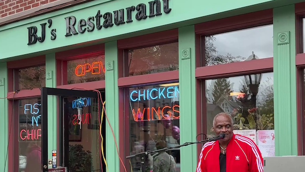 a man plays music in front of the green exterior of BJ's Restaurant with "BJ's Restaurant" written on the front.